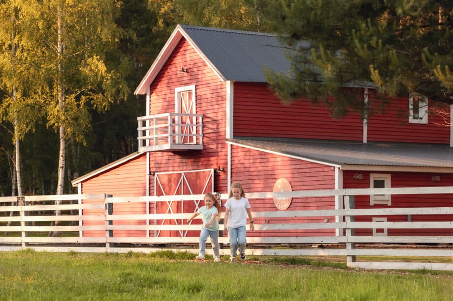 A red barn behind a white fence with two girls nearby