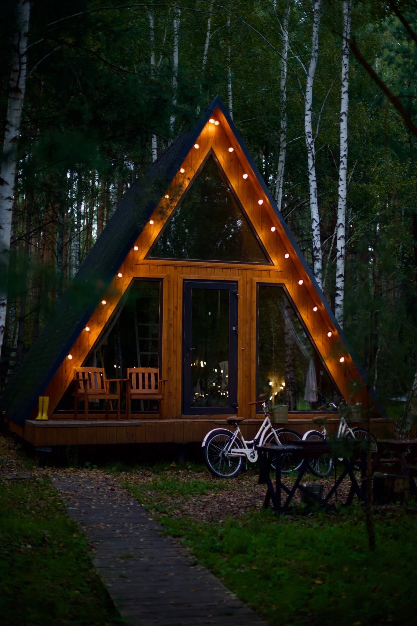 An A‑frame cabin with string lights among birches in the evening, bikes by the porch